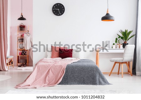 Pink blanket on bed next to desk and grey chair in bedroom interior with black lamp and clock