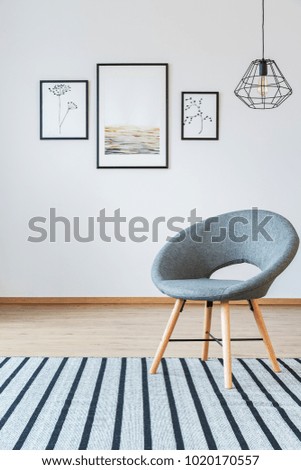 Grey armchair on striped carpet in simple living room interior with gallery of posters and lamp