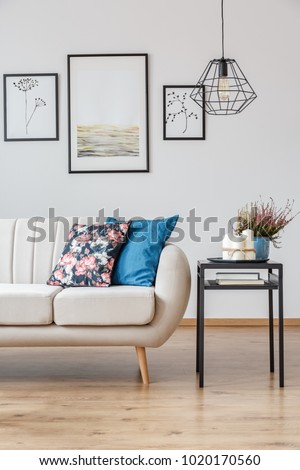 Lamp above plant and candles on table next to sofa with floral pillow in living room interior with posters