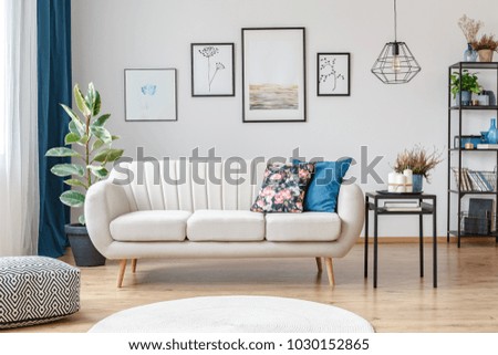 Floral pillow on beige sofa next to table with candles in living room interior with pouf and posters