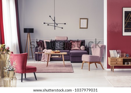 Pink and red armchair in warm living room interior with pillows on settee against the wall with poster