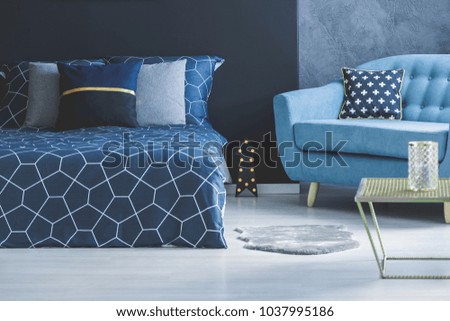 Blue sofa with patterned cushion and grey fur next to a dark bed in cozy bedroom interior with table