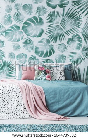 Decorative cushions with plain and floral prints on a turquoise bed by a leaves wallpaper in bedroom interior