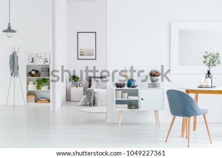 Grey chair at wooden table in white open space interior with poster on the wall and bright bedroom