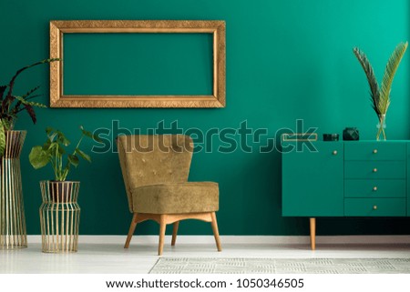Palm leaf on a modern, teal sideboard with drawers in a luxurious, green living room interior with golden decorations and an upholstered chair