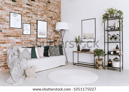 Posters on red brick wall above white sofa with pillows in bright living room interior with plants