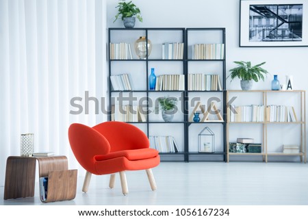 Orange armchair next to wooden table in spacious living room interior with bookshelf and poster