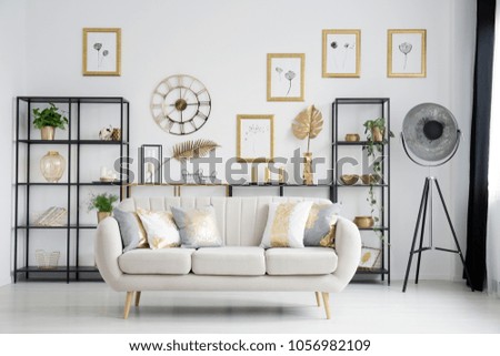 Beige sofa with gold cushions in bright living room interior with industrial lamp and posters