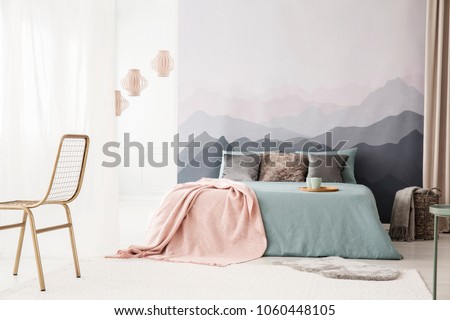 Tray with mugs on a cozy, blue bed with a pastel pink blanket by a landscape wallpaper in a bright bedroom interior