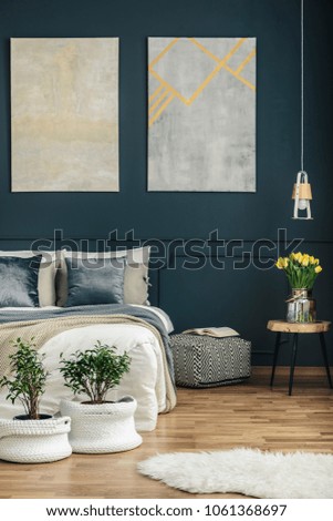 An open book on an ottoman standing by a dark blue wall between a bed and a stool with yellow tulips in a modern bedroom interior