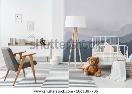 Grey armchair and wooden lamp on carpet in child's bedroom interior with plush toy and mountain wallpaper
