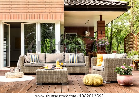 Yellow pouf next to a rattan armchair and flowers on wooden patio with striped pillows on sofa