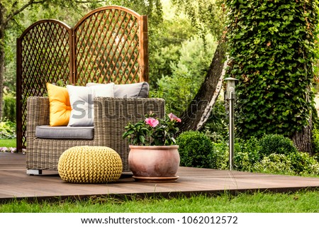 Yellow pouf next to a rattan armchair with grey and gold pillows and pink flowers on wooden veranda