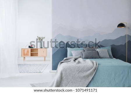 Blue bed with grey blanket against mountain wallpaper in simple bedroom interior with wooden cupboard