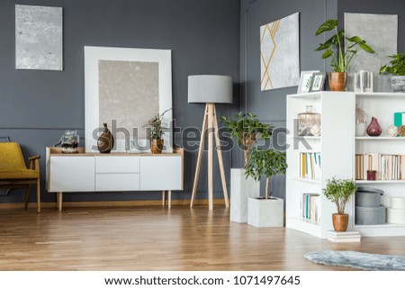 Books on white bookshelf and plants in floral grey living room interior with paintings on the wall