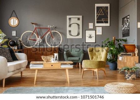 Art collection on a grey wall, wooden coffee table, armchair and red bike in a messy living room interior