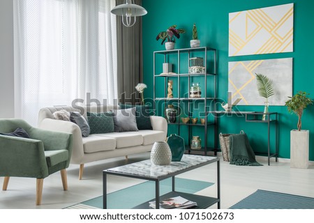 Bright sofa with many pillows standing next to a green armchair in living room interior with metal furniture and geometric paintings