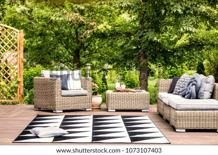 Pillow on geometric carpet between rattan garden furniture against green tree during summer