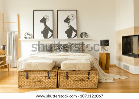 African posters, wicker boxes, fireplace and double bed with patterned pillows in a boho bedroom interior
