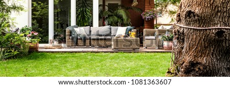Wicker sofa with pillows and table with fruits and juice standing on garden terrace by the house with glass door