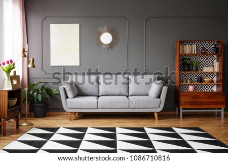 Black and white carpet with geometric pattern placed on the floor in dark living room interior with grey couch, vintage cupboard with books and wainscoting on the wall