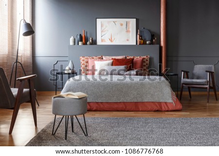 Real photo of cozy, dark bedroom interior with many decorative cushions on the bed with gray blanket and vintage armchairs