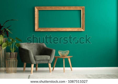 Wooden table with gold, metal bowl standing by a green armchair in living room interior with frame on the wall