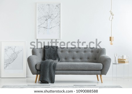Grey lounge with dark blanket standing in white living room interior with two map posters, gold bulb lamp and books on a small table