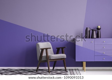 Real photo of a gray, wooden armchair on patterned, black and white rug in creative living room interior with geometric, violet wall and cupboard