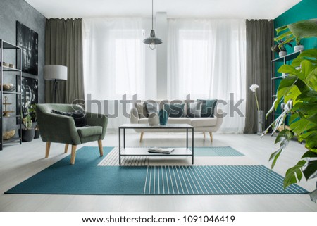 Table on carpet next to a green armchair and beige sofa in bright living room interior with plant