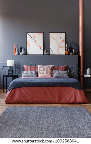 Front view of a cozy bed with ginger bedding, gray blanket and pillows in dark bedroom interior with black bedside tables, watercolor posters and elegant decorations. Real photo
