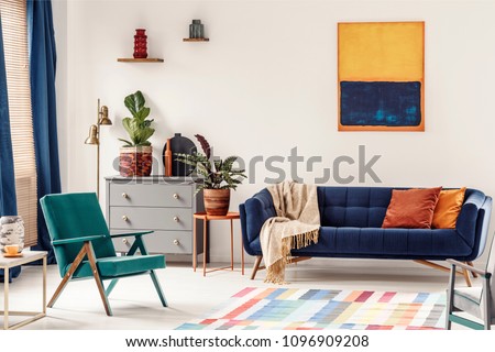 Orange end table with fresh plant standing next to navy couch with blanket and pillows in white living room interior with painting, green armchair, vases on shelves and colorful carpet on the floor