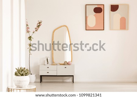 Plant on table and mirror on white cabinet in simple living room interior with posters. Real photo. Place for your furniture