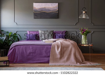 Lamp above copper table with flowers next to bed with violet bedding in purple bedroom interior
