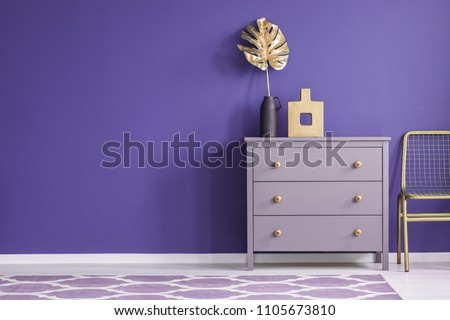 Gold vase on violet cabinet next to a chair in purple living room interior with patterned carpet