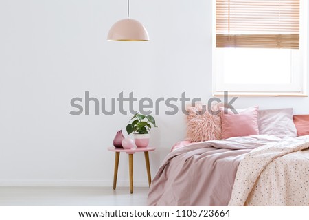 Cozy, feminine bedroom with pink bed, decorative cushions and plant on a wooden stool standing against white, empty wall. Real photo with a place for your furniture.