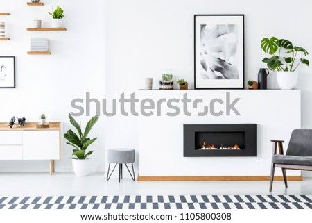 Grey armchair next to fireplace under poster in living room interior with plant and stool. Real photo