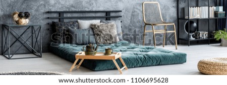 Gold chair placed in dark bedroom interior with raw wall, green mattress bed with pillows and books on metal rack