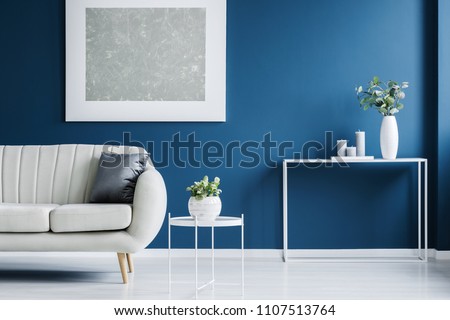 Metal console table with plant in vase and candles standing against blue wall in living room interior with light grey sofa and modern poster