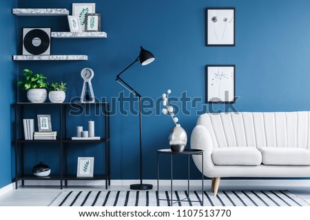 Fresh green plants, candles and books placed on a black metal rack in blue living room interior with simple posters and bright sofa