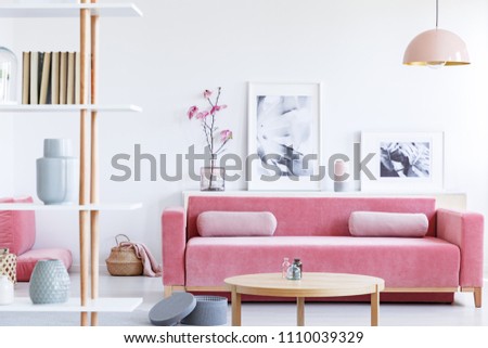 Posters and flowers above pink sofa in pastel living room interior with wooden table. Real photo