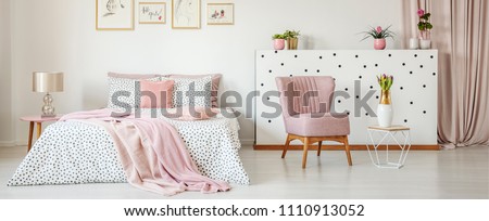Dotted bedding on double bed placed in white room interior with green plants, fresh tulips, simple posters and pastel pink armchair