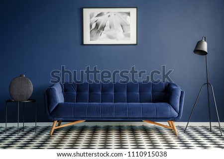Navy blue room interior with comfortable plush couch in the middle, black lamp and side table with decoration standing on chessboard floor. Framed image on the wall. Real photo