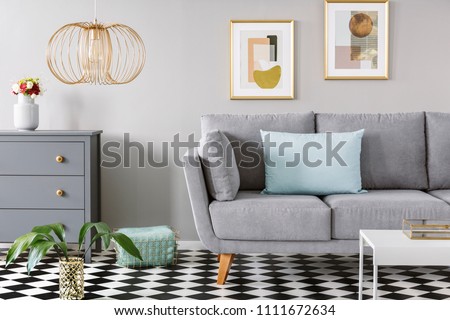 Light blue pillow placed on grey couch in bright living room interior with checkerboard linoleum floor, fresh flowers in vase on cupboard, two posters hanging on the wall and gold lamp