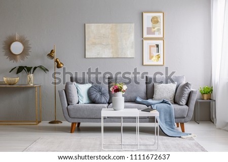 Grey sofa with pillows and blanket standing in bright living room interior with gold lamp, fresh flowers on white table and carpet on the floor