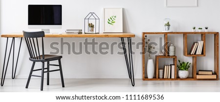 Hairpin table with computer, notebooks, poster and cactus standing in white home office interior with books and plants on wooden shelves