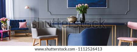 Chairs standing by the blue and gold dining table with pink roses in vase in dark grey open space living room interior