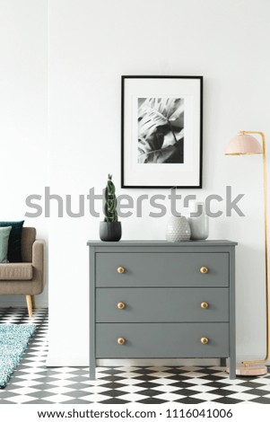 Black and white poster hanging on the wall above grey cupboard with decor standing in white room interior with checkerboard linoleum floor