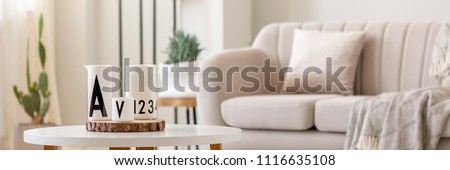 Close-up photo of white vessels standing on white end table in bright living room interior with sofa and plants in blurred background