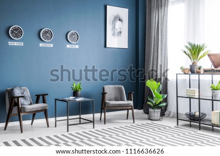 Two grey armchairs, metal rack with decor, window with drapes and plants placed in dark living room interior